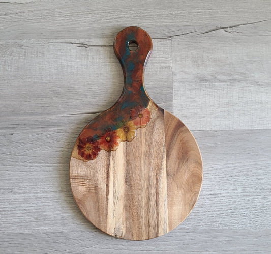 3-D Flowers are captured in resin on this paddle style cutting board. Flowers in shades of orange, burgundy and yellow. The resin on the board is in sparkling teal, dark orange and a little bit of gold. The paddle board has a hole for hanging. Care: Occasionally treat the wood with Olive Oil or Food Grade Mineral Oil. Clean the resin with a damp cloth or an alcohol wipe. Approximate size: 16 x 10 inches.