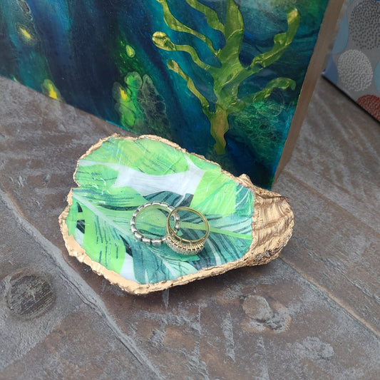 Hand Crafted Tropical Leaf design has a coating of Resin and Gold Trim with a coat of varnish. Shades of Green on white background. Gold Trim. Perfect little touch to any bedside or bathroom. Brighten an entry hall or even use as a salt server. (Food Safe) Use alone as a pretty accent to a TV stand, shelf or bookcase. The possibilities are endless! Resin is UV and Food Safe. Approximate size at longest and widest points: 4 x 3 inches Rings NOT included