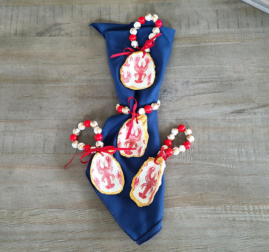 Set a beautiful, unique table with these lobster themed napkin rings. They can also be used to decorate a bottle, vase or door handle. The oyster shells are covered with a lobster design and trimmed in gold. Each shell is coated with an acrylic spray. I hand painted each wood bead. White with gold beads and red beads. Each one is tied with a red silk ribbon. Napkin shown is NOT included.