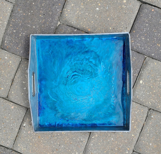 This metal tray is versatile. Use it for holding your remotes, serving drinks or just let it sit on your cabinet and gaze at the depths formed in the resin. Colors of shimmering blues, teals and pewter make up the design. Square Metal tray has cut out handles on two ends. Approximate size: 10.5 x 10.5 x 2 Inches
