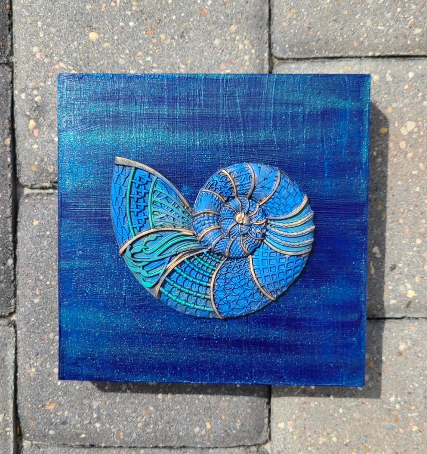 I created this very detailed Nautilus out of a resin mold. The beautiful colors really show off the detail. I painted the wood background with shimmering blues and teals. The whole canvas sparkles! This piece is so unique and detailed. Truly one-of-a-kind. Saw tooth hanger on the back. Ready to hang or can stand alone on a shelf. Lightweight Approximate size: 6 x 6 x 1.25 inches.