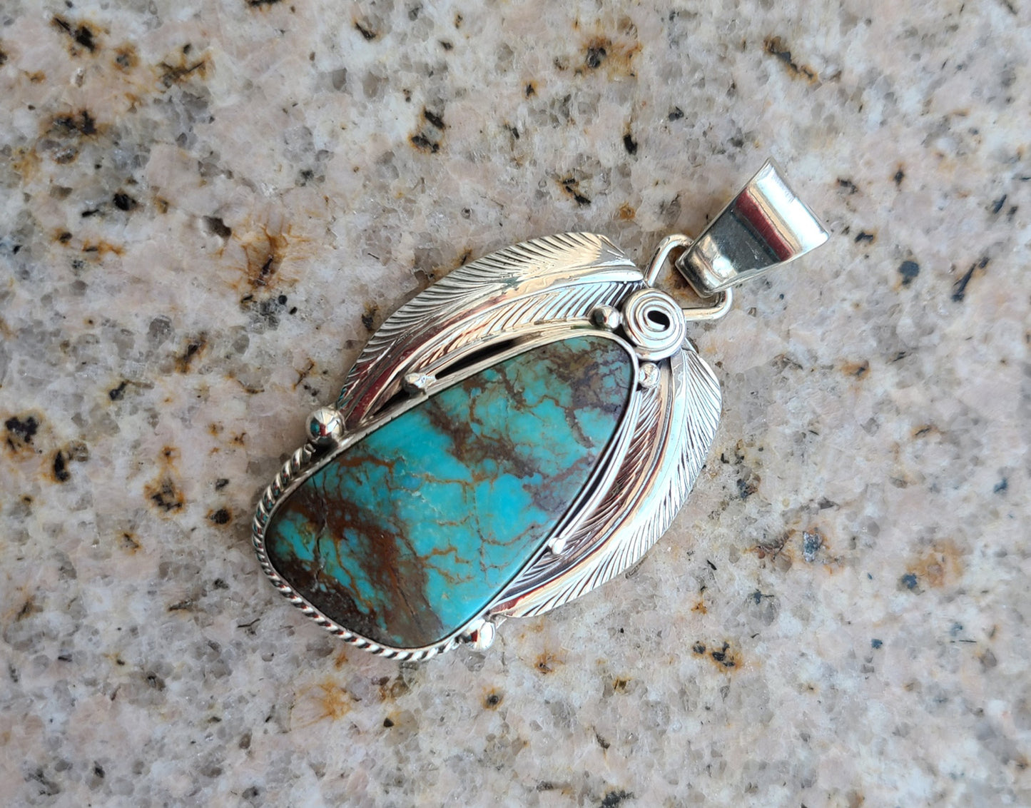 Large freeform Kingman Turquoise has various shades of blue and green with black and brown matrix. The stone is accented with a large silver feather on each side, and twisted rope on the bottom part. Navajo artist, Davey Smith. Approximately 2.25 inches long.