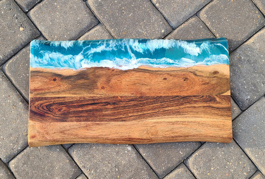 Large cutting or charcuterie board with live edge on both the long sides. An angry ocean, turquoise blue sparkles beneath white caps of waves. Resin is UV resistant and food safe. Resin covers approximately 2.5 inches of one long edge. Overall size is approximately 20 inches long and 10.75 inches wide. Acacia Wood. Hand wipe and treat with food grade mineral oil or olive oil. Resin can be cleaned with an alcohol wipe.