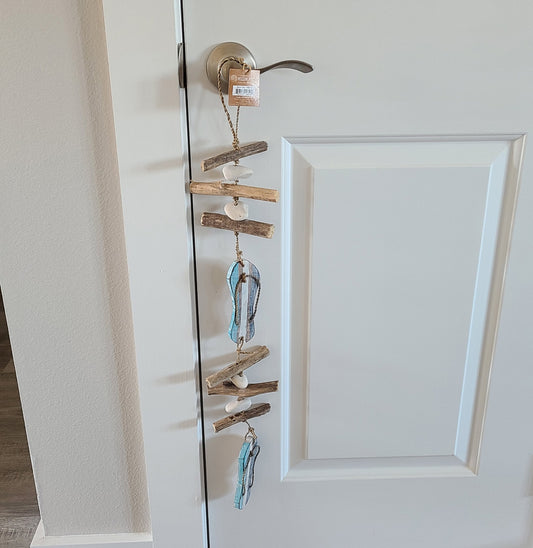 Sticks of wood with stones between them. Two wooden flip flops hang in the middle and on the end. All connected by jute rope with a loop for hanging. Overall length is approximately 27 inches.