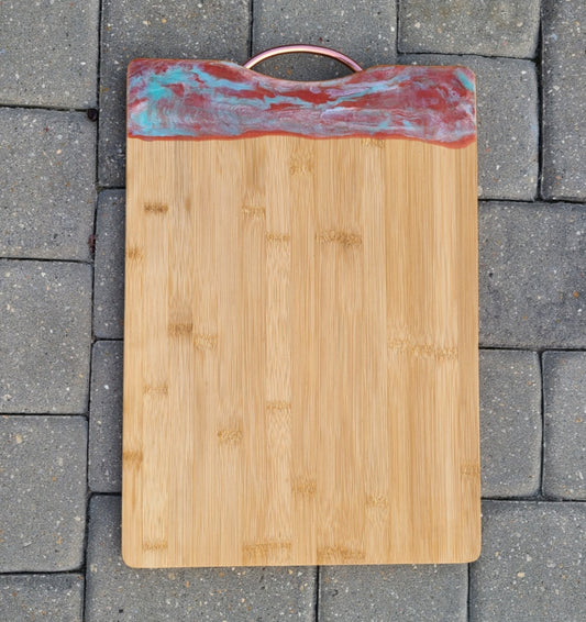 Shades of copper, rust, teal and pearl white resin float on this bamboo cutting board. Resin is UV Resistant and Food Safe. Approximate size: 17.75 x 12.5 inches; the handle adds another 1.25 inches to the overall length of the board. To clean the resin, wipe with a damp cloth or an alcohol wipe. Board weighs about 3 lb 12 oz.