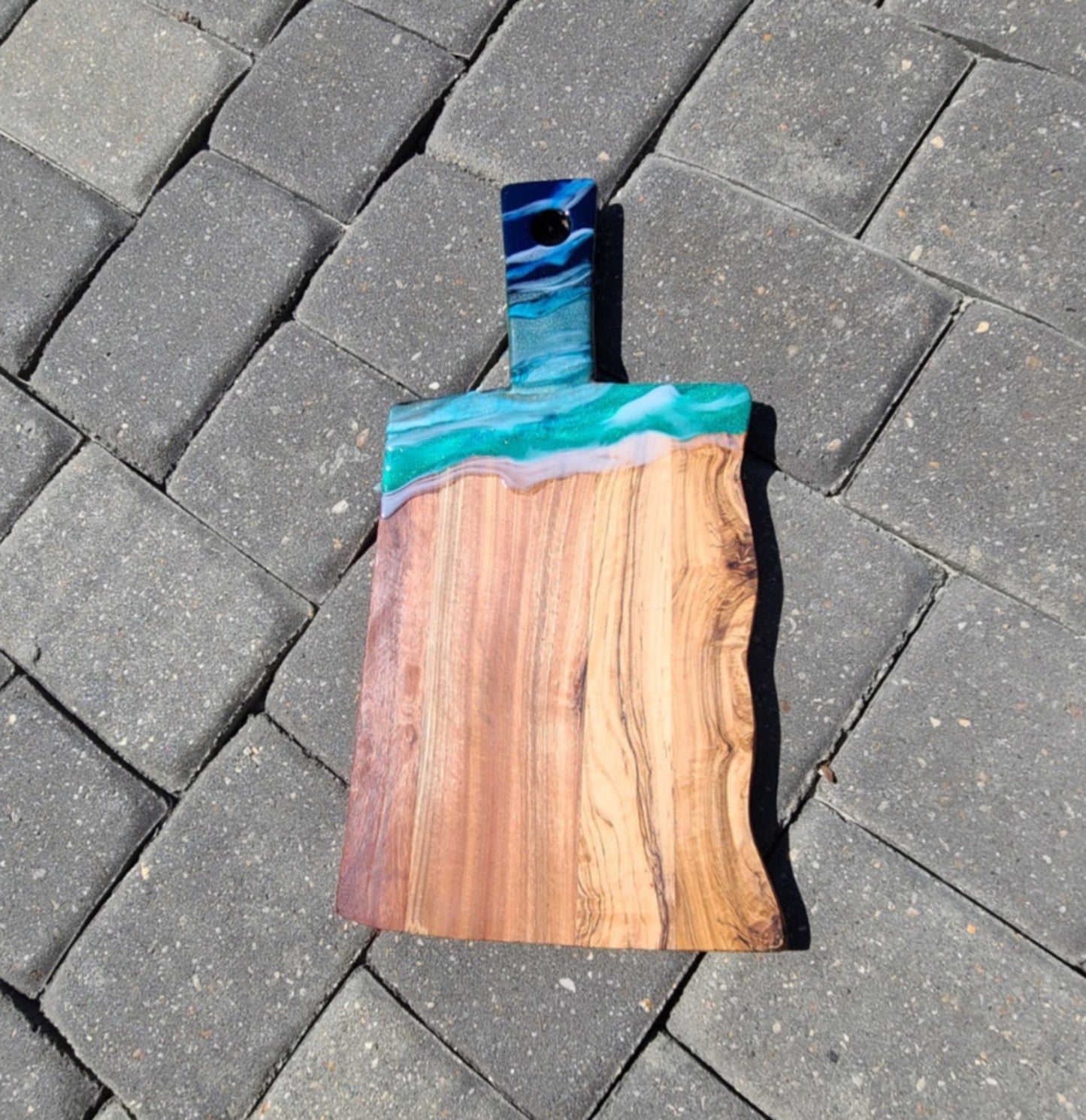Beautiful board. Olive Wood Cutting/Charcuterie Board with one live edge. I accented the top portion with food grade resin. Lots of sparkling blues and green dance below the white waves. Nice thick, heavy board. Approximate size: 16.75 x 9 inches Care: Hand wash and dry thoroughly. Occasionally, treat the wood with food grade mineral oil. The resin can be wiped clean with a damp cloth or an alcohol wipe.