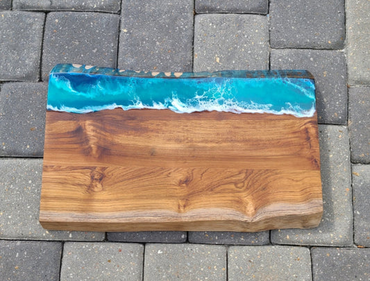 This cutting board or charcuterie board is 1.25 inches thick with a live edge and a beautiful resin ocean scene. Deep dark blues give way to teals and of course, the white waves. Resin is UV Resistant and Food Safe I believe the wood is Acacia Approximate measurements: 18 x 10-11 inches. 1.25 inches thick. Heavy board weighs about 5.5 pounds!