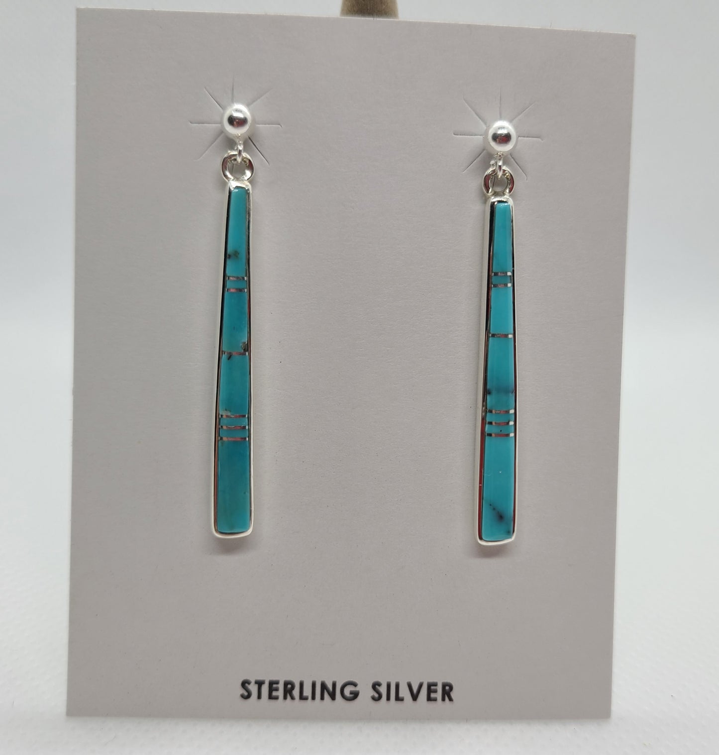 1.5 inch long earrings by Navajo Artist, Rick Tolino. Inlay Turquoise are bright blue with a small amount of black matrix. Sterling Silver ball studs have a small loop that the earrings dangle from. They are like a long stick with small pieces of Sterling between the Turquoise.