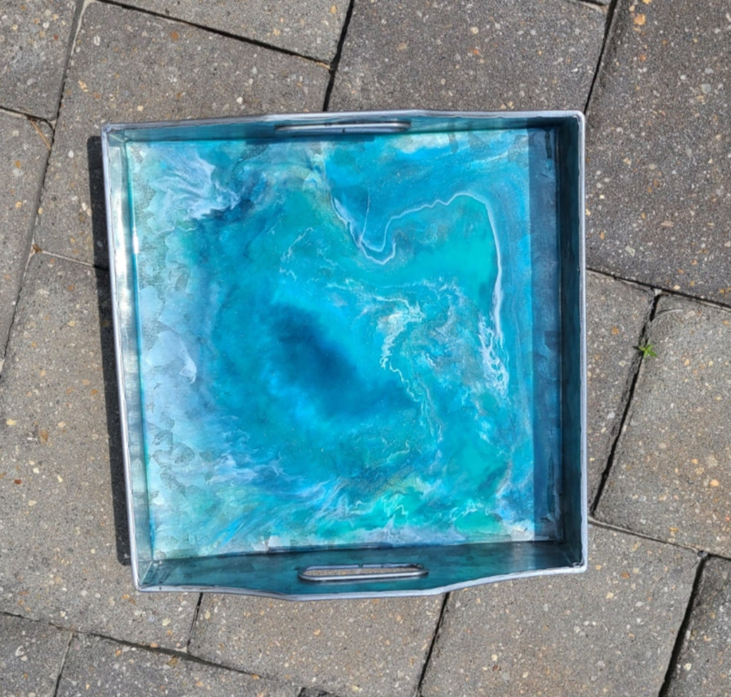 This metal tray has a resin design that looks like a deep blue vortex. Lightweight metal tray looks like it is made of Aluminum and has cutouts for handles on two sides. Use as serveware or as a trinket dish. Would be perfect for holding remotes on the coffee table. Resin is UV resistant and food safe. Approximate size: 9.5 x 9.5 inches square and 1.75 inches deep on the sides without the handles. Care: Wipe with a damp cloth or clean resin with an alcohol wipe.