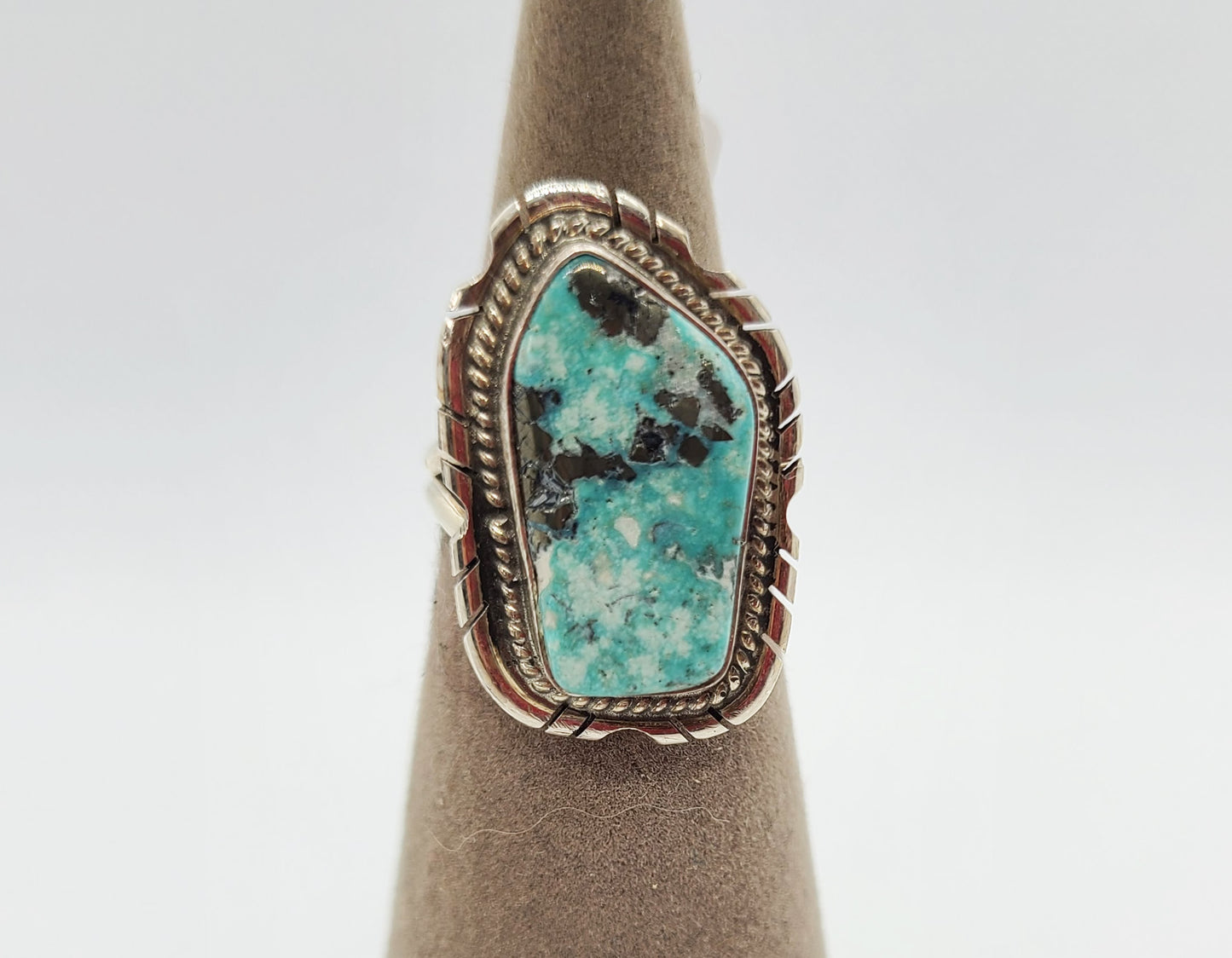 Freeform shape Kingman stone is approximately .75 inches long. Flat on one end and a rounded point on the other. Robin's egg blue with silver matrix and speckles of white. Bezel set with twisted wire and saw-cuts on the backplate. It has a split band. Size 6. Navajo artist Peggy Skeets. Stamped Sterling and P. Skeets. MSRP is $125