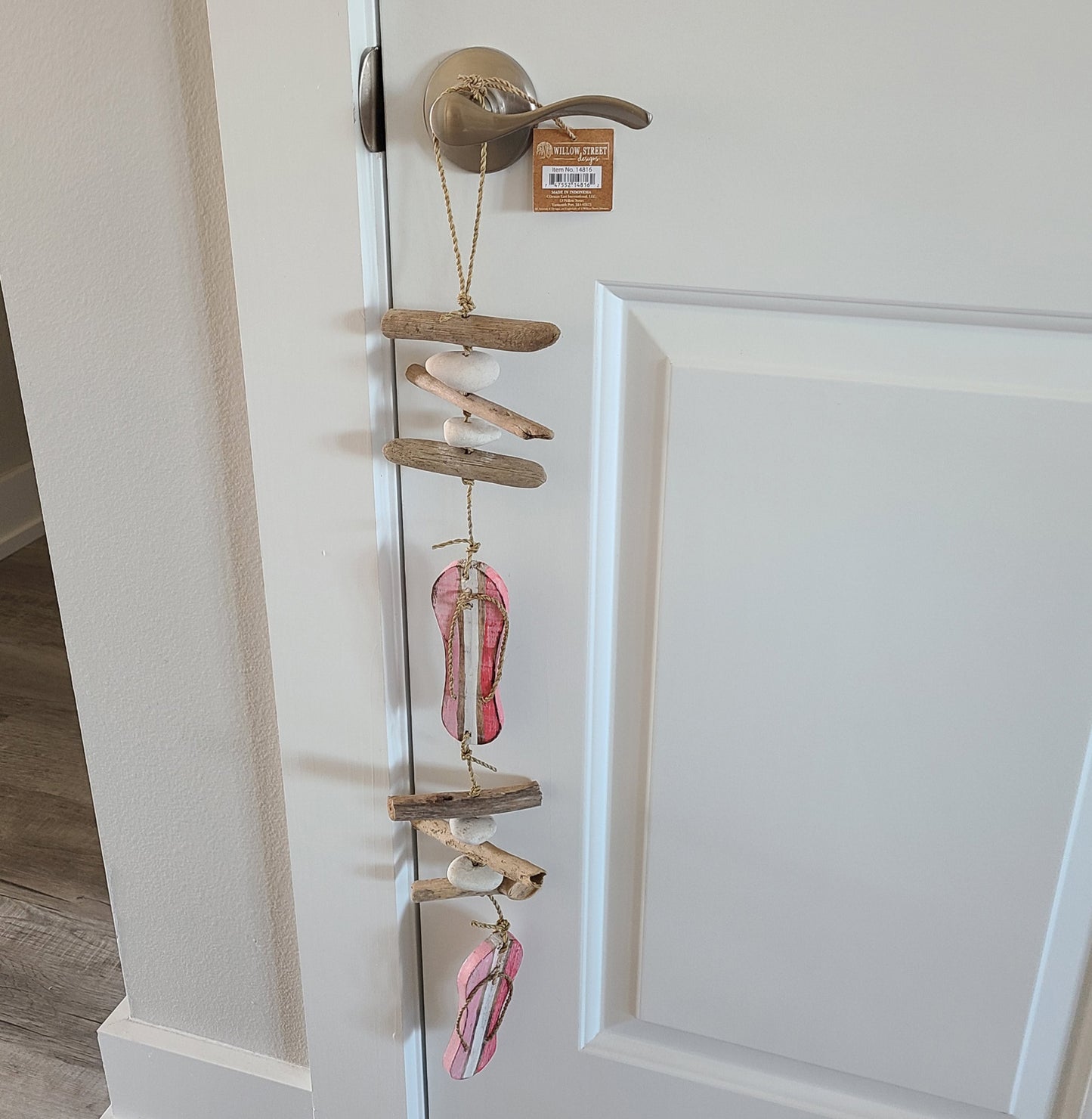 Two wood cut out flip flops are pink with white stripes. Six pieces of round wood are separated by white stones. All are hung on a jute rope and the top has a big loop for hanging. Great for indoors or out. Overall length is approximately 27 inches.