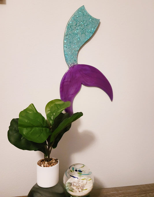 MDF Board is shaped like a mermaid tail. The body portion is blue green with sparkles of chunky glitter and the tail is a bright purple. A small amount of crushed glass separates the tail from the body. The entire piece is covered in UV resistant resin. Saw tooth hanger on the back. The approximate size is 19 x 9 inches.