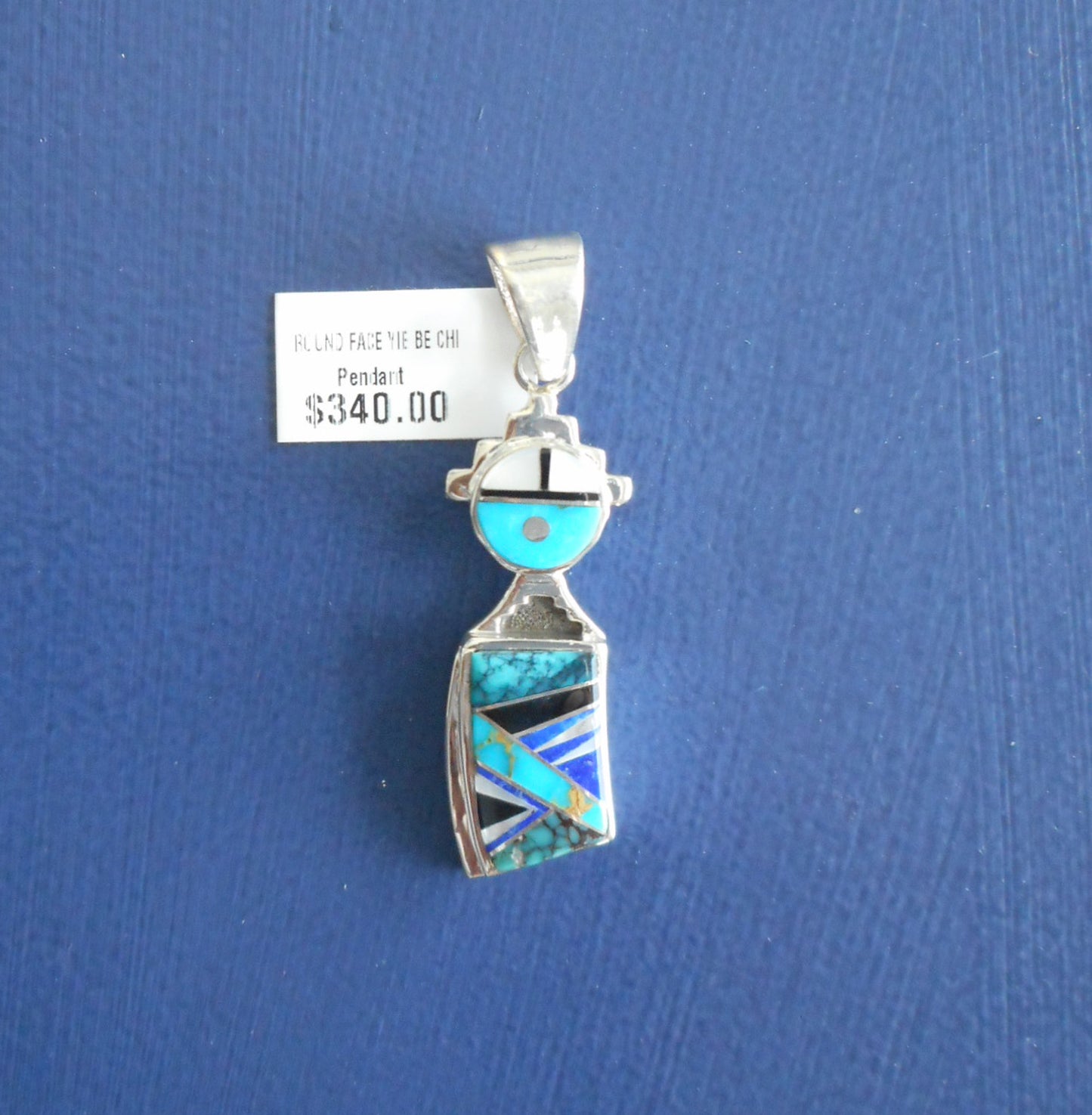 Round faced Yei pendant is made of inlaid stones of turquoise, only, lapis and Mother of Pearl. Cut out silver around the head and on the neck. Silver bail. 2 inches long including the bail and .50 inches wide.