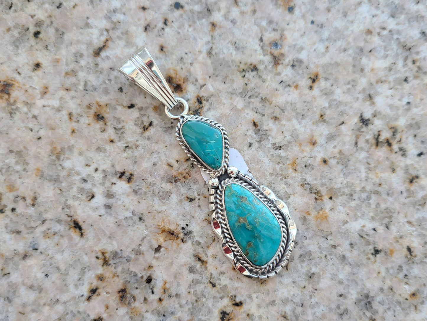 Two stone turquoise pendant. Kingman, AZ turquoise. Navajo artist Sam Yellowhair. Set in Sterling Silver. Small twisted wire around the smaller top stone and twisted wire around the larger bottom stone with back-plate that has cut-outs on it. Approximately 2.5 inches long including bail. Bail is large enough to accommodate most necklaces.