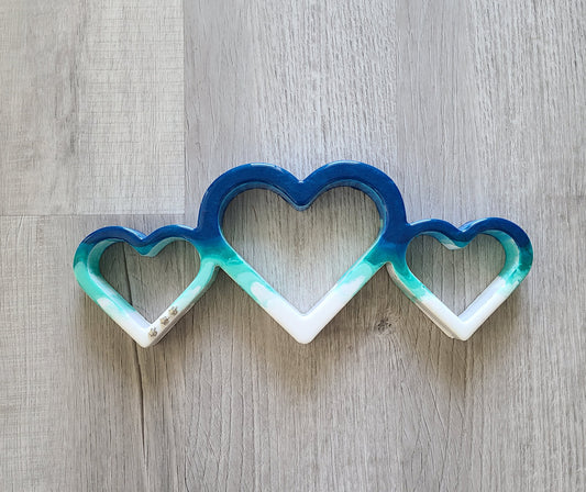 The three hearts connect to the ocean and features three little turtles racing toward the water.  One of a kind piece.  UV Resistant resin on wood  Turtles are metal  Stand or hang  Approximate measurements: 14.5 x 6 x 1.25 inches