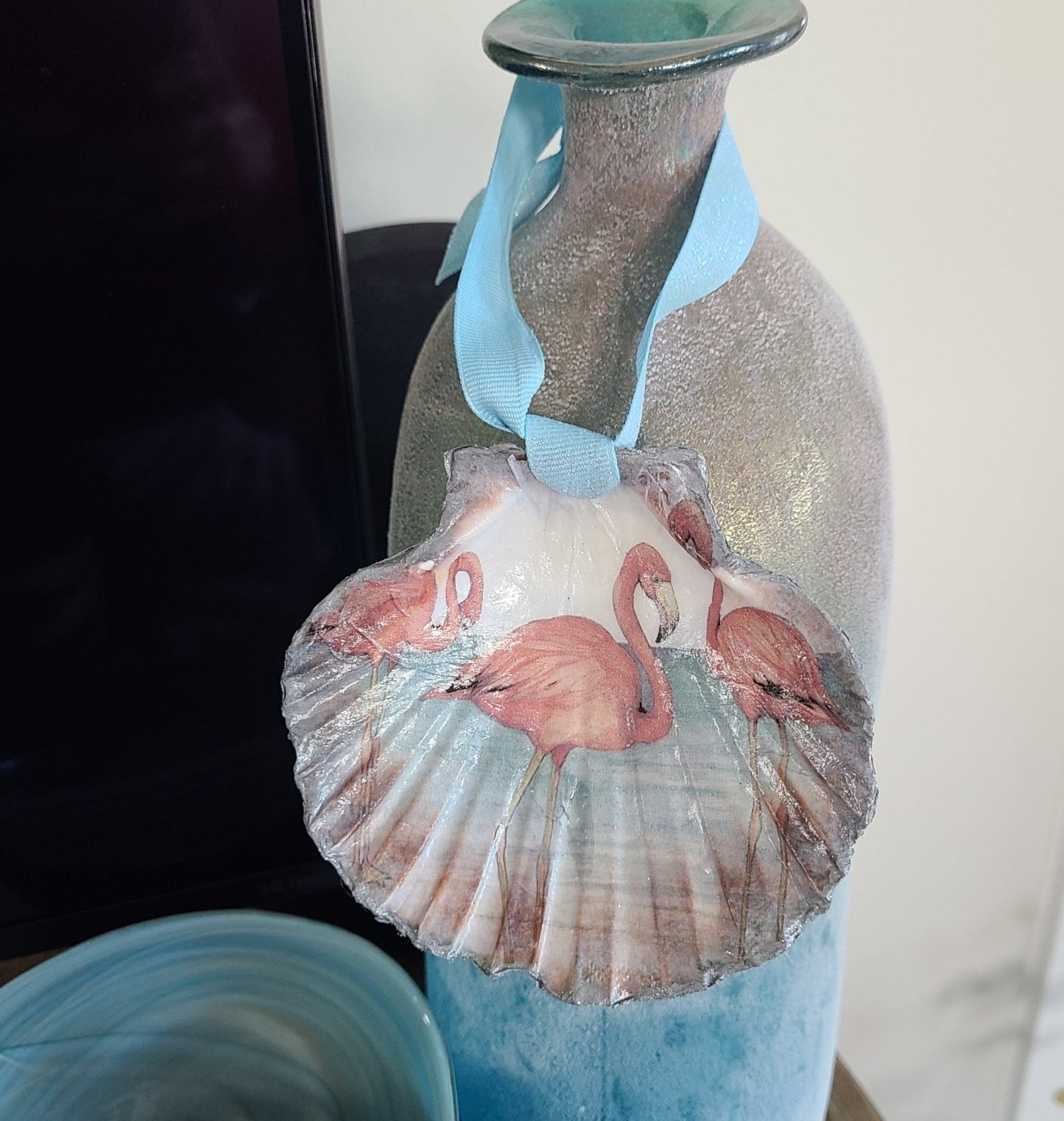 I decorated this clam shell with a design of three flamingos.  The back side looks like wave ripples.  The edges are trimmed in metallic silver.  A blue iridescent ribbon is tied on for hanging.  Acessories shown are NOT included.  Approximate size: Shell: 4 x 4.25 inches