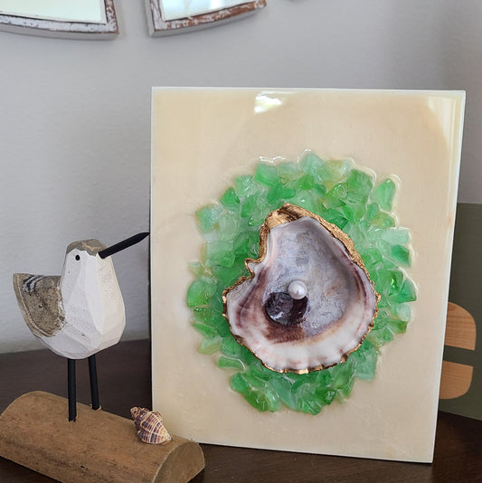 This is a beautiful real oyster shell showing off her beauty in this artwork.  The shell is trimmed in gold and coated with varnish.  A single pearl is nestled in the middle.  Crushed green glass surrounds the shell and lies on a soft beige background.  This piece will stand alone or you can hang it on the wall.  Resin is UV Resistant  Approximate size: 8 x 6.25 x 1.75 inches  Accessories shown in the lifestyle photos are NOT included.