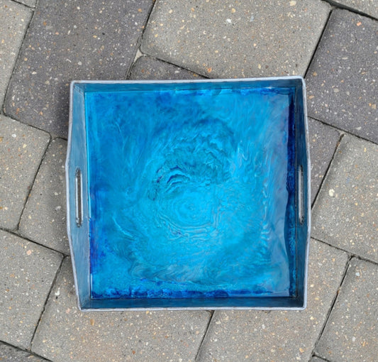 This metal tray is versatile. Use it for holding your remotes, serving drinks or just let it sit on your cabinet and gaze at the depths formed in the resin.  Colors of shimmering blues, teals and pewter make up the design.  Square Metal tray has cut out handles on two ends.  Approximate size: 10.5 x 10.5 x 2 Inches