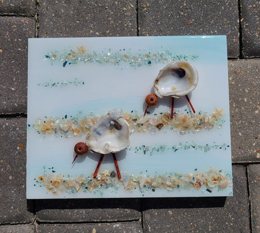 Two cute little shorebirds peck along the shore, looking for treasures.  I created this artwork on wood using seafood green crushed glass, their heads are wooden beads and stick legs and beak, oyster shells and shells. The shells are from Panama City Beach. The entire piece is covered in UV Resistant resin.  One-of-a-kind piece of artwork for your walls.  Approximate size: 8 x 10 inches