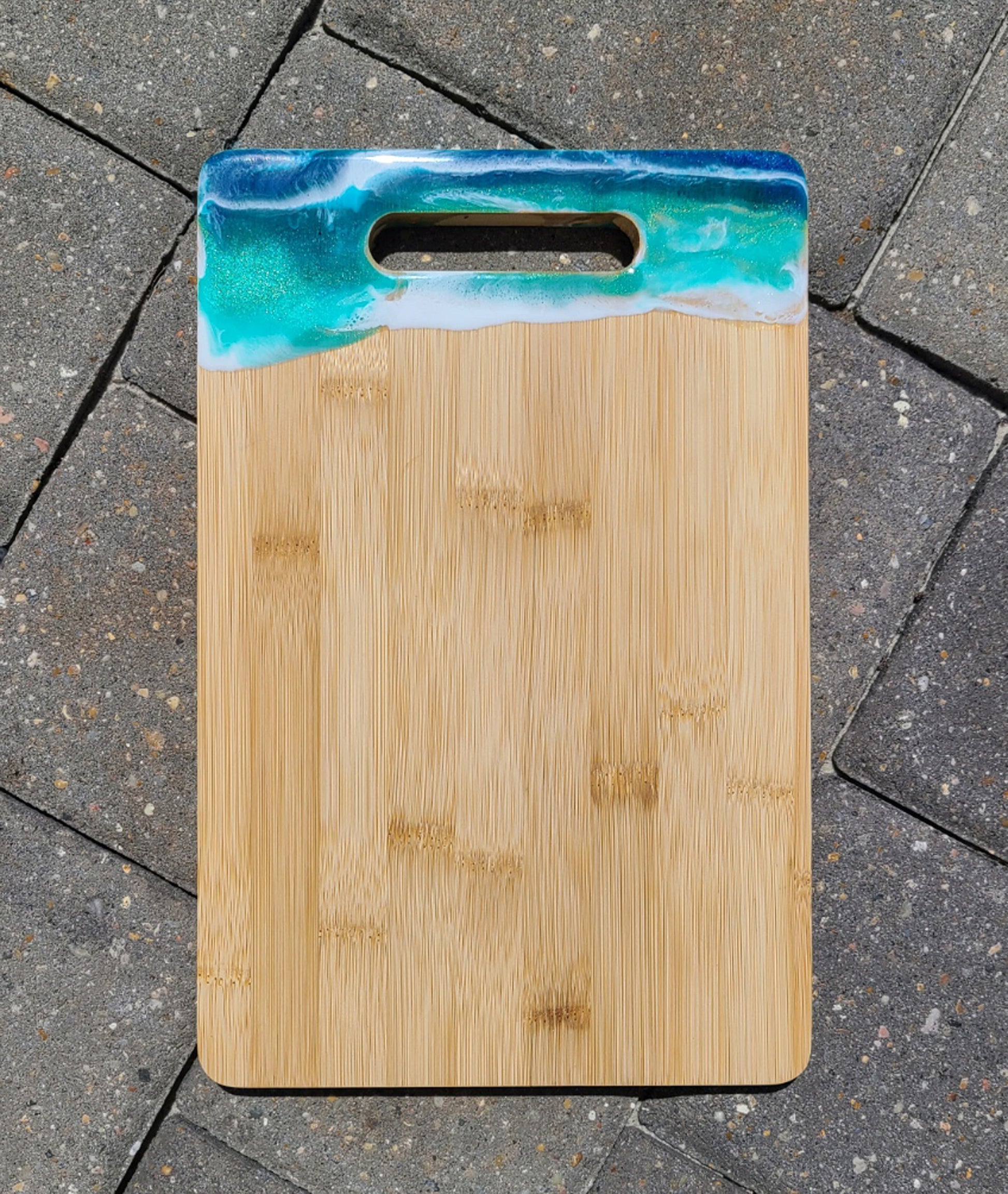 Sparkling colors of the ocean and white waves give this cutting board a touch of serenity for any home.  Functional as well as beautiful.  Bamboo board by Brookstone.  Approximate size: 8 x 12 inch  Resin is UV Resistant and Food Safe.  Care: Hand Wash Warm Water, Wipe with a damp cloth or alcohol wipe.