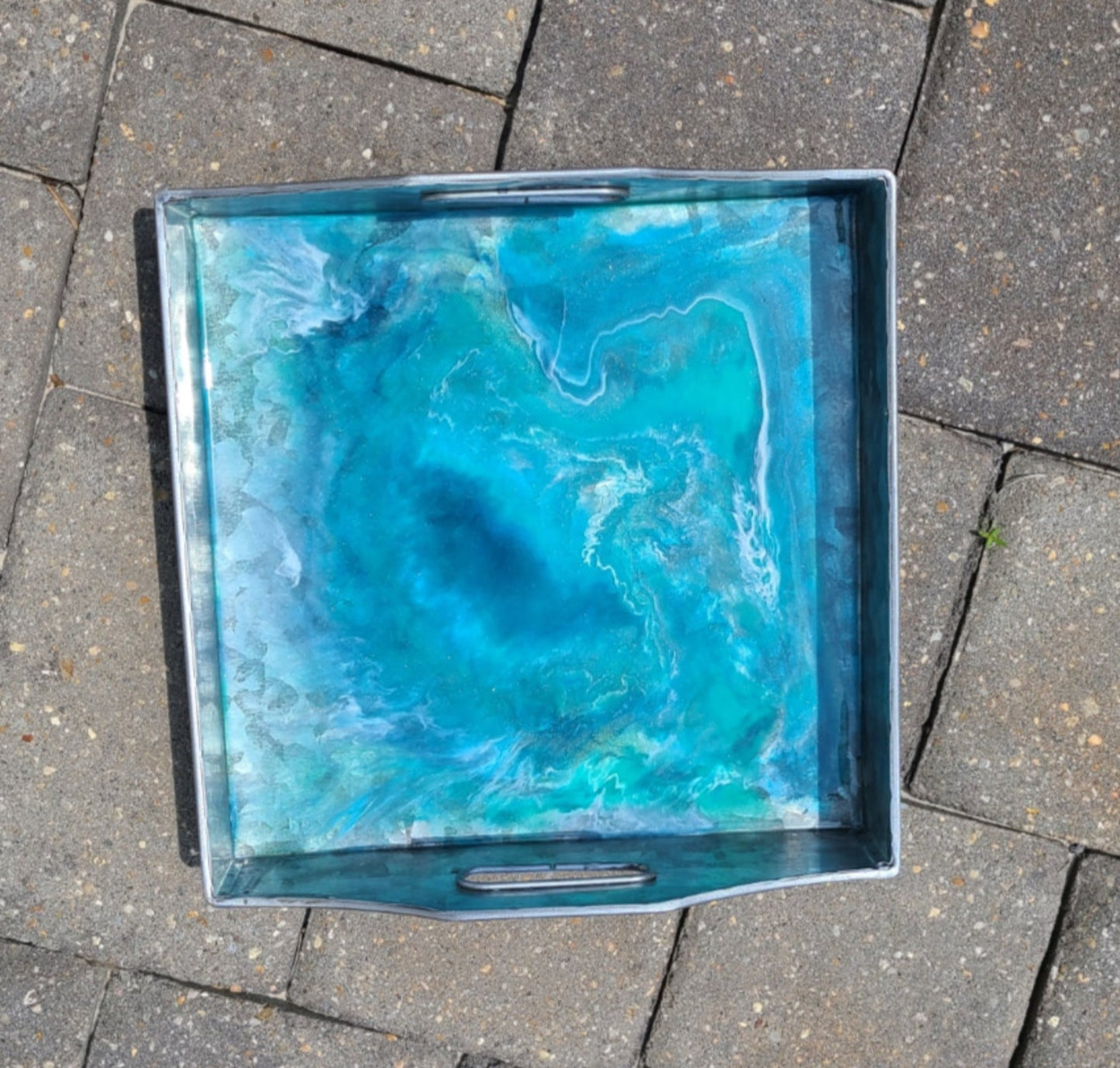 This metal tray has a resin design that looks like a deep blue vortex.  Lightweight metal tray looks like it is made of Aluminum and has cutouts for handles on two sides. Use as serveware or as a trinket dish. Would be perfect for holding remotes on the coffee table.  Resin is UV resistant and food safe.  Approximate size: 9.5 x 9.5 inches square and 1.75 inches deep on the sides without the handles.  Care: Wipe with a damp cloth or clean resin with an alcohol wipe.