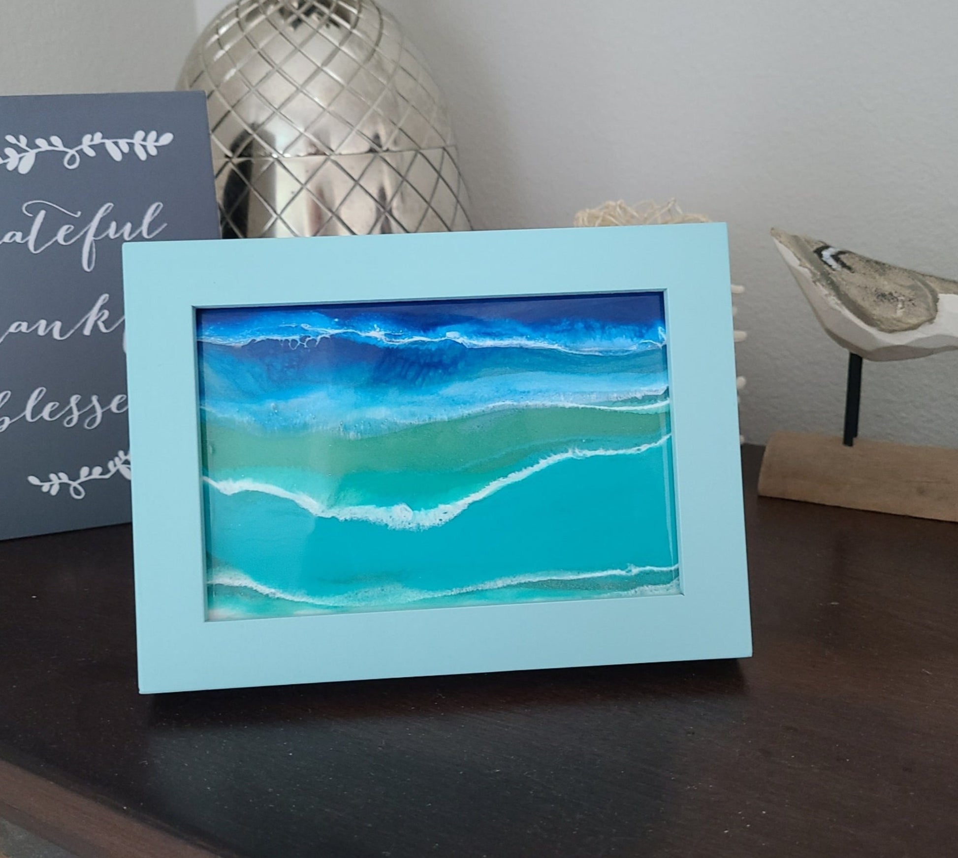 Need a cute picture in a small spot? I had so much fun making these ocean scenes in frames. This one is in a sea foam blue/green wood frame.  When you touch and feel the "picture", it feels like glass.  I took the glass from the frame and resined an ocean scene on it.    The frame can stand alone or be hung.  Approximate size:  7.25 x 5.25 inches overall; inside dimensions are approximately 5.5 x 3.75 inches.