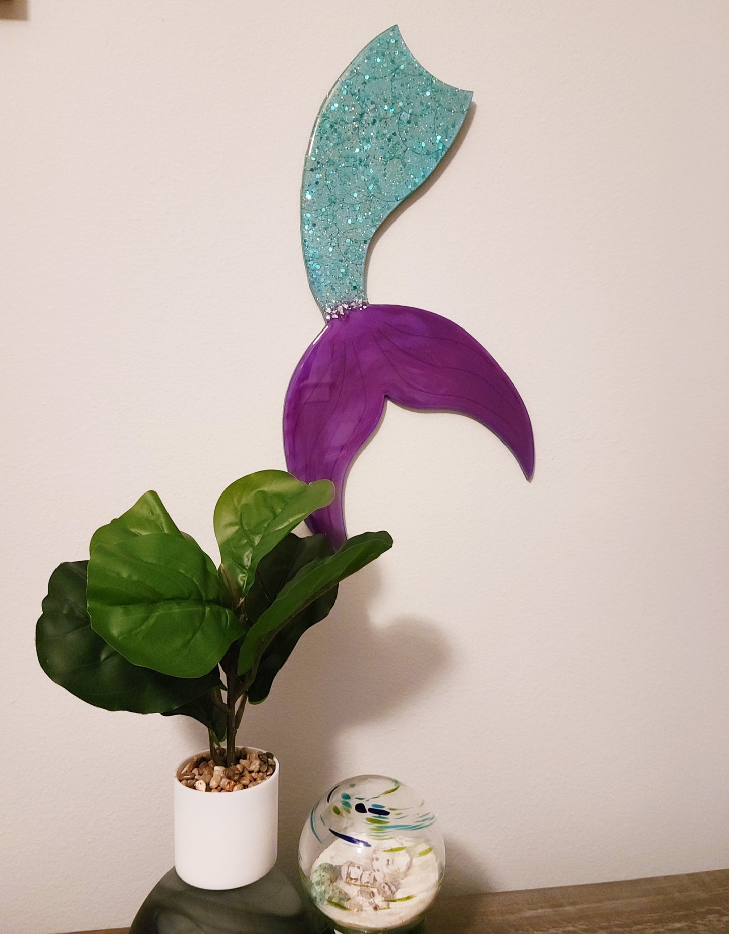 MDF Board is shaped like a mermaid tail.  The body portion is blue green with sparkles of chunky glitter and the tail is a bright purple.  A small amount of crushed glass separates the tail from the body.  The entire piece is covered in UV resistant resin.  Saw tooth hanger on the back. The approximate size is 19 x 9 inches.