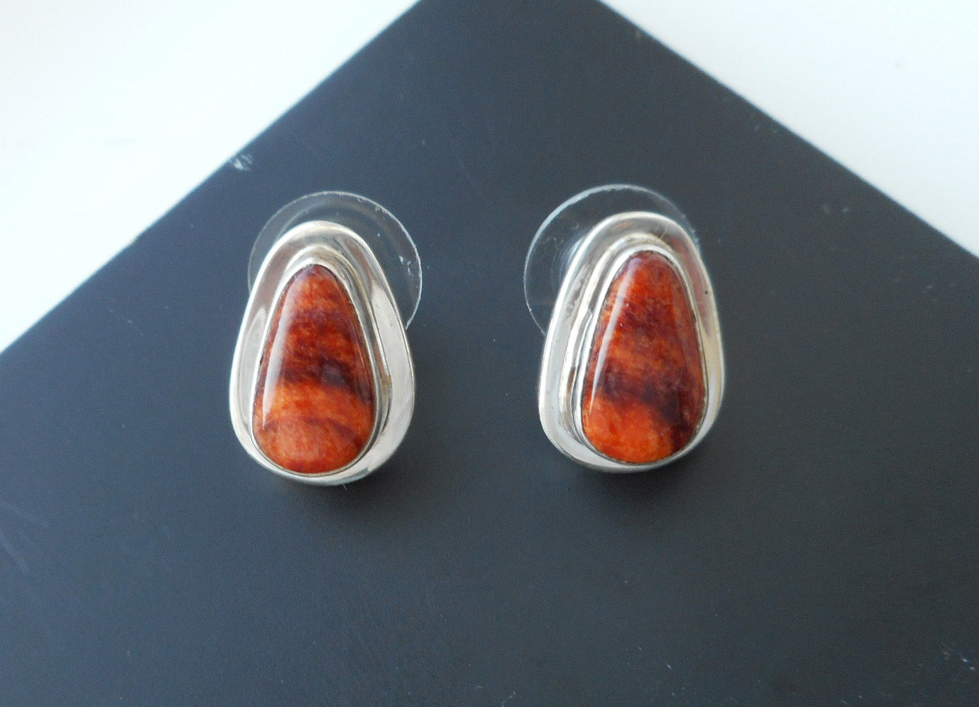 Stud Earrings. Beautiful Orange Spiny Oyster stones reminiscent of a fiery sunset.  Bright and dark orange has subtle bands of black.  The stones are a soft teardrop shape.  Set in Sterling Silver bezel style with a small amount of backplate showing on the edge. Overall size is approximate 5/8 inch.  